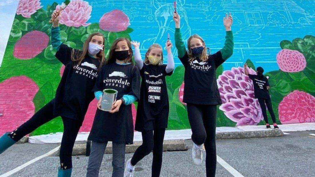  Group of young girls in "Cloverdale" sweatshirts posing joyfully in front of a vibrant mural with floral designs, celebrating community spirit.
