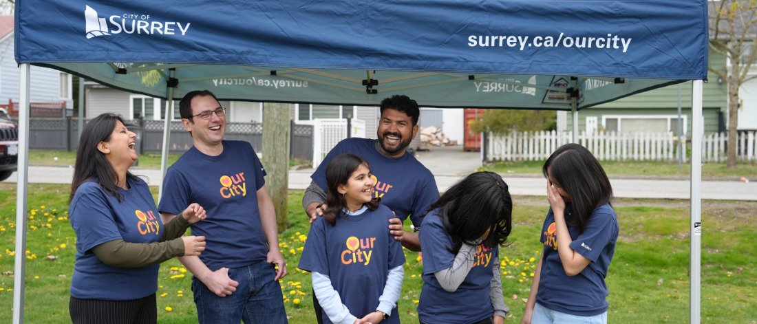  A joyful group of people wearing "Our City" t-shirts under a City of Surrey tent, sharing a laugh together at a community event.
